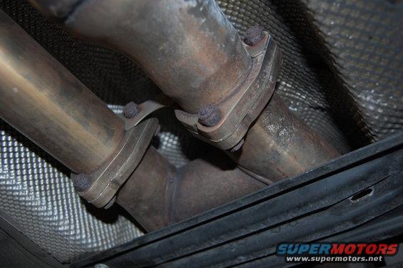 Close-up of exhaust pipes connecting under a vehicle. There's a Supermotors.net logo in the corner