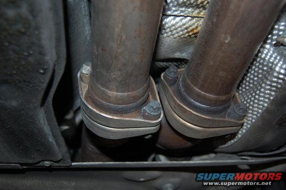 Exhaust pipe flanges and bolts on a vehicle underside, with Supermotors.net logo visible