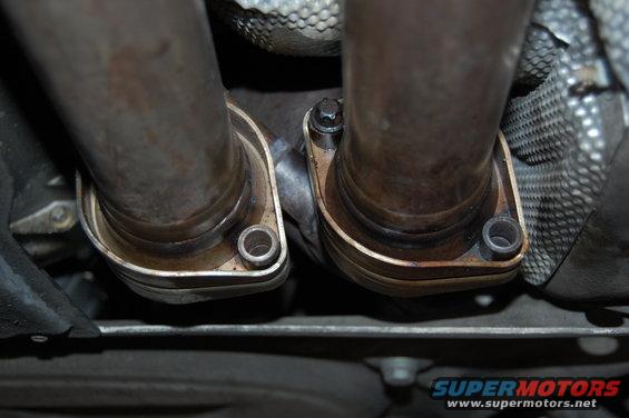 Close-up of two exhaust pipes connecting to flanges under a vehicle. A Supermotors.net logo is visible at the bottom right