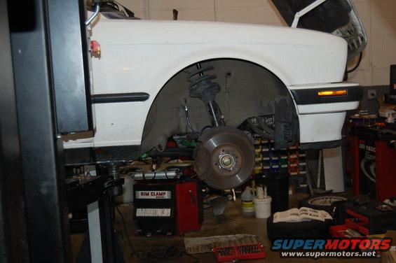 White car on lift, front wheel removed, showing suspension and brake rotor. Orange turn signal visible
