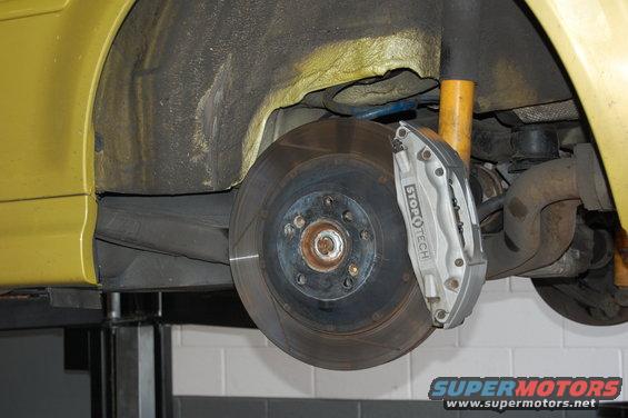 Close-up of a car's front wheel assembly, showing a StopTech brake caliper and rotor. Yellow body panel visible