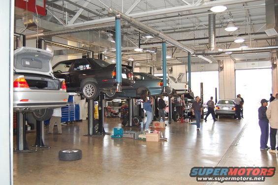 Silver BMW M3 on a lift, trunk open, in a garage with other cars and people