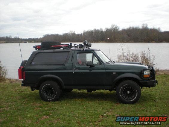 Dark green 1993 Ford Bronco with roof rack and spare tire parked on grass near a body of water