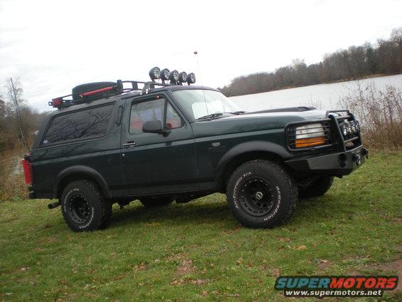 Dark green SUV with roof rack, spare tire, and brush guard parked on grassy terrain near water