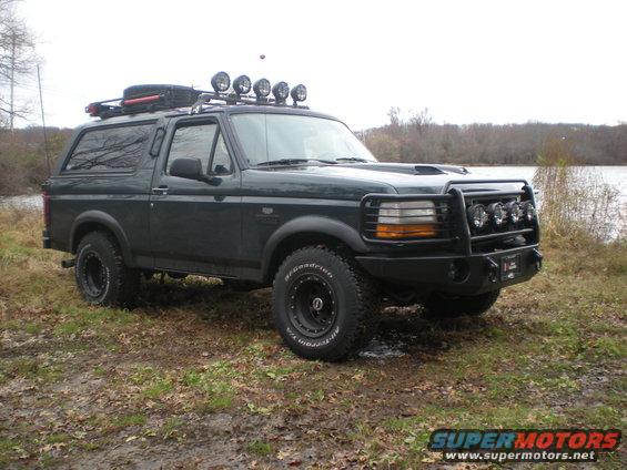 Dark green 1993 Ford Bronco with roof rack, light bar, and brush guard parked on muddy ground near water