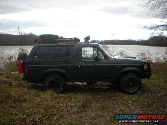 Dark green Ford Bronco parked by a lake, with a spare tire and gas can mounted on the back, and gear on the roof rack