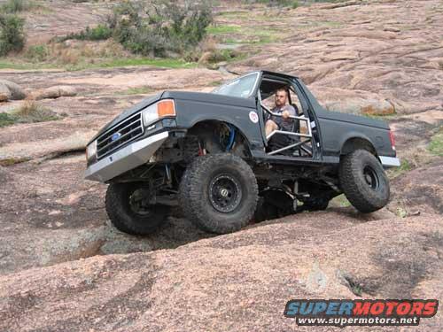 Ford pickup truck with lifted suspension and large tires climbing over rocks. Driver is visible inside