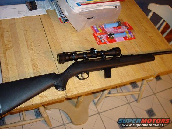 Black rifle with scope on a wooden table, next to PEZ dispensers and papers