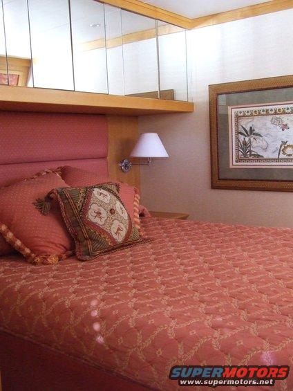 Bedroom with a made bed, pillows, a lamp, and framed art on the wall. Mirrors are above the headboard