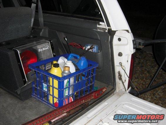 Interior cargo area of a vehicle showing a blue crate of cleaning supplies, a subwoofer box, and a Hi-Lift jack