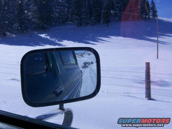 Green vehicle side mirror view of snowy road with multiple vehicles behind