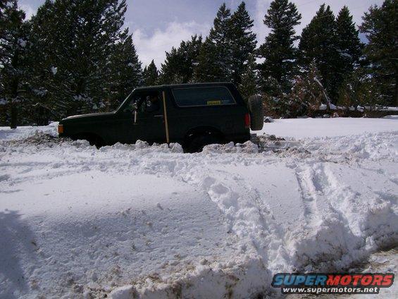 Dark green SUV with spare tire on rear stuck in deep snow, shovel visible in driver's door