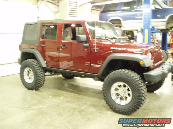 Red Jeep Wrangler Rubicon with aftermarket wheels and tires in a garage. Another vehicle is visible on a lift in the background