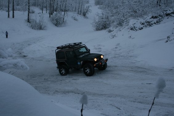 Jeep Wrangler with roof rack driving on a snowy road, trees in background