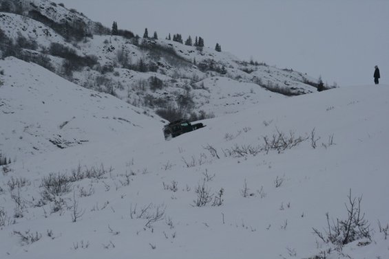 Dark SUV navigating a snow-covered hillside with a person standing on a ridge above