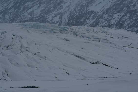 Vast, snow-covered glacier with textured ice formations and a distant, snow-dusted mountain range under an overcast sky
