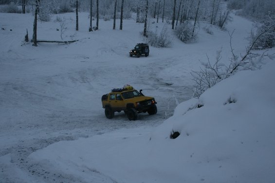 Yellow off-road SUV with roof rack and snorkel driving in snow, another dark SUV behind it