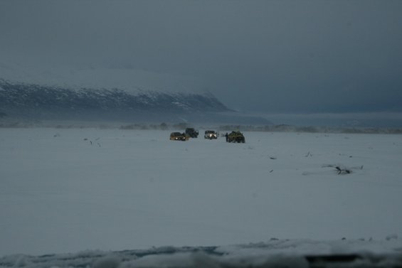 Several vehicles drive across a vast, snow-covered plain with mountains in the background under a cloudy sky