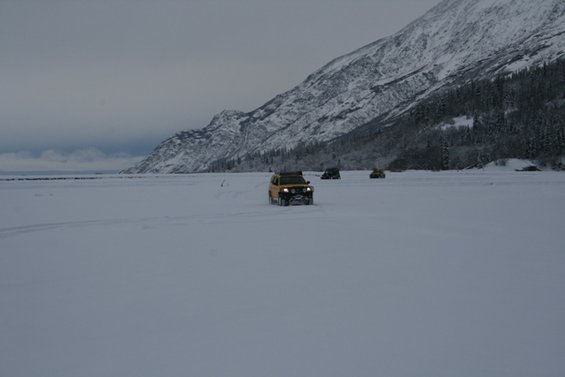 Yellow SUV and two other vehicles driving across a snowy plain with mountains in the background