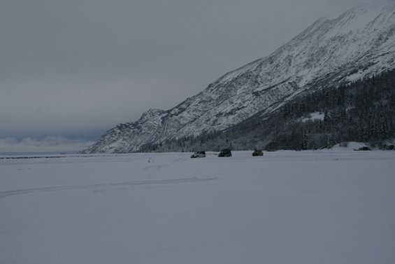 Three SUVs drive across a vast, snow-covered plain towards a , snow-capped mountain range under a cloudy sky