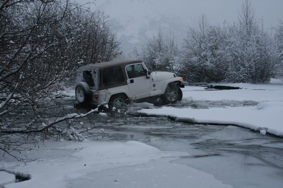 White Jeep Wrangler driving through icy water with snow-covered trees in background