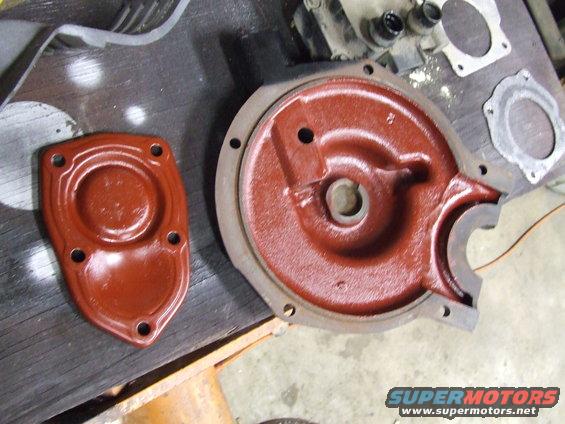 Two red-painted metal engine parts on a wooden surface. One is a flat cover with four bolt holes, the other is a larger, circular housing with a cut-out section