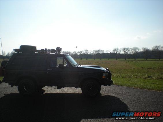Black Ford Bronco parked on asphalt with a field and trees in the background. It has a roof rack with a spare tire