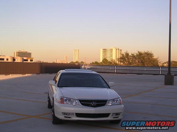 White sedan parked on a rooftop parking lot, front view. The car has a sunroof and aftermarket wheels
