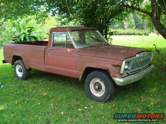 Rusty red pickup truck parked on grass under a tree. Visible Jeep grille and chrome bumper