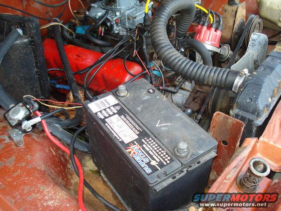 Engine bay with a black battery, red engine block, carburetor, and hoses. Rusty metal surfaces are visible