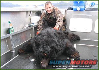 Man in camo jacket poses with a large black bear on a boat deck. Water and mountains in background