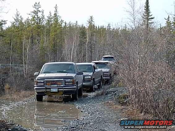 Front view of a 1999 GMC Sierra 1500 driving through a muddy trail with other trucks behind it
