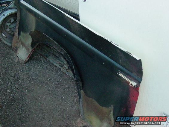 Damaged dark-colored vehicle fender with rust showing, next to a wheel and tire
