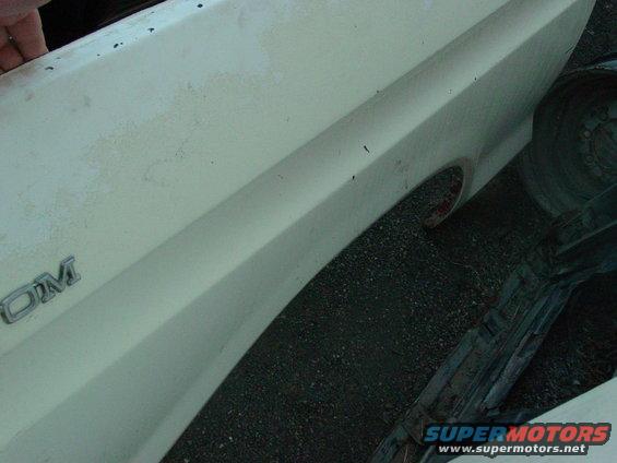 Close-up of a white vehicle fender with a chrome emblem that reads 'OM'. The wheel well and part of a tire are visible