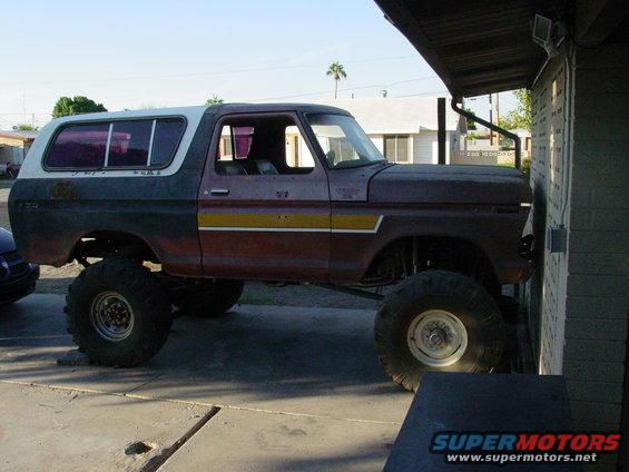 Lifted 1978 Ford Bronco with white camper shell, maroon and dark gray body, yellow stripe, parked against a brick wall