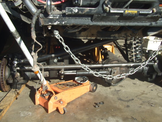 Underside of off-road vehicle with suspension components, differential, and a chain. Orange hydraulic jack is positioned under the frame