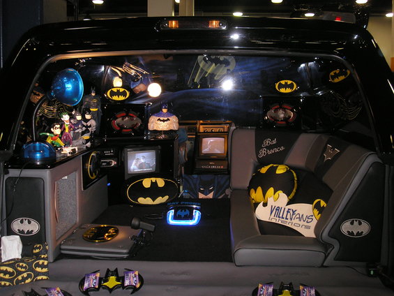 Interior of a vehicle customized with Batman memorabilia, including figures, pillows, and screens. Text on seat reads 'Bat Bronco'