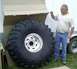 1990 Ford Bronco - A man stands beside a massive tire, dwarfed by its size, with a trailer in the background.