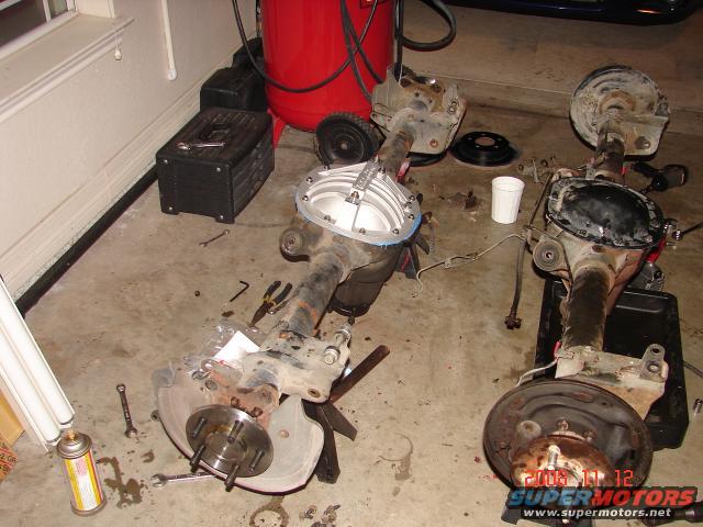 Two disassembled car rear axles on a concrete floor with tools and a red air compressor