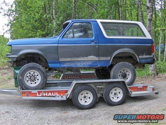 1990 Ford Bronco on a U-Haul auto transport trailer, side view. The Bronco has a blue and gray two-tone paint scheme