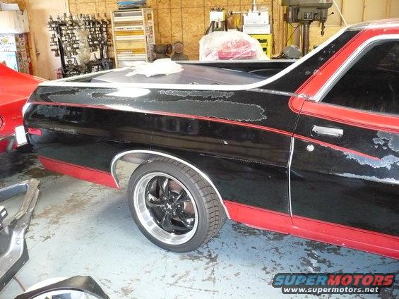 Black and red Ford Ranchero in a garage, paint peeling on the side. Another red vehicle is partially visible
