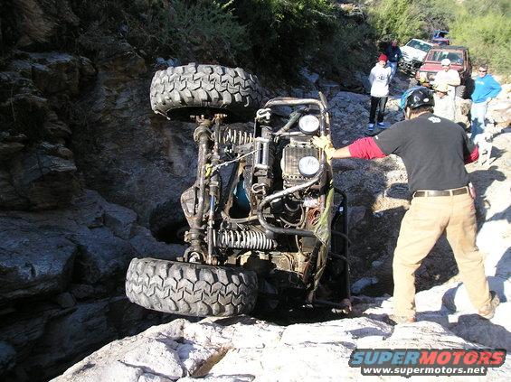 Off-road vehicle on its side with people around, other vehicles in background