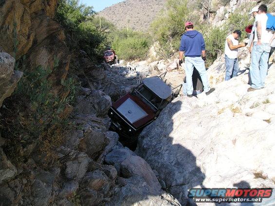 Two off-road vehicles are stuck in a narrow, rocky canyon. People are standing around observing