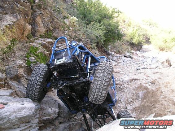 Blue off-road buggy with large tires is tilted on its side, climbing over large rocks on a steep, dry, mountainous trail