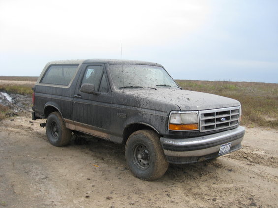Mud-covered Ford Bronco parked on a dirt road with marshland in background