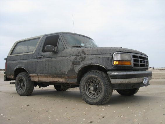 Mud-covered dark gray 1992 Ford Bronco on a sandy beach, viewed from the front three-quarter angle