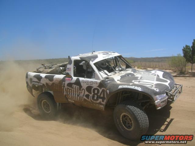 White and black cow-patterned off-road truck with number 84, kicking up dust on a dirt track
