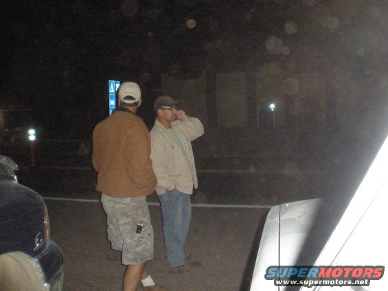 Two men stand on a street at night near a parked car