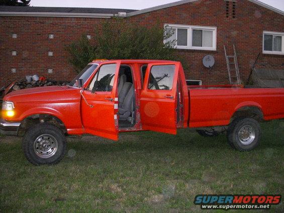 Red crew cab pickup truck with all doors open, parked on grass in front of a brick house