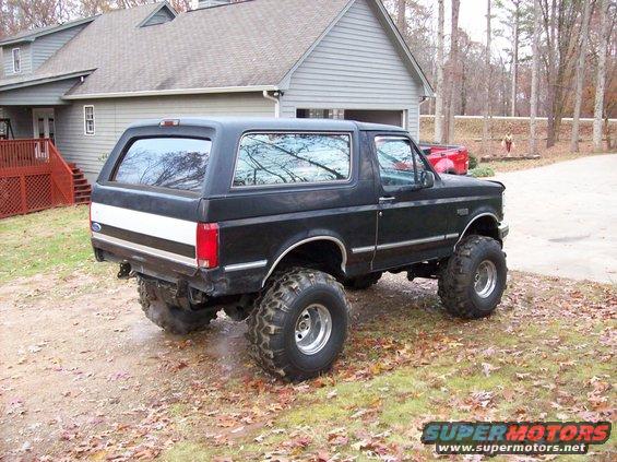 Black lifted Ford Bronco parked in front of a house. It has large off-road tires and a white stripe on the rear